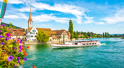 A scenic view of Stein am Rhein, Switzerland, featuring a picturesque riverside town with colorful historic buildings and a tall church steeple. A white tour boat cruises along the sparkling blue waters of the Rhine River. Bright flowers in the foreground frame the scene, while the clear blue sky with wispy clouds enhances the vibrant atmosphere