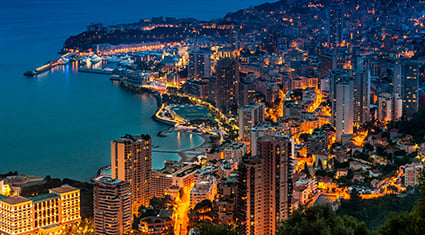 A stunning nighttime view of Monte Carlo, Monaco, illuminated with vibrant city lights. The image showcases the luxurious coastline, high-rise buildings, and winding streets, with the Mediterranean Sea glistening in the background. The dramatic hillside setting adds depth and grandeur to the scene, reflecting the city's opulence and charm.