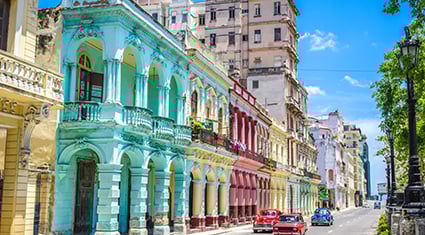 Colorful colonial buildings line a sunny street in Havana, Cuba, with classic American cars driving by under a bright blue sky.