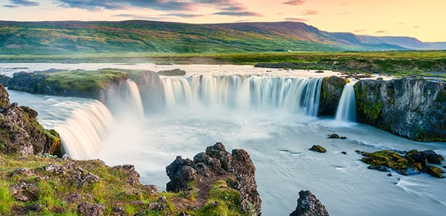 A breathtaking view of Goðafoss waterfall in Iceland, with powerful cascades of water flowing over a wide cliff into a calm river below. The lush green landscape surrounding the waterfall contrasts with the rugged rocks and the soft hues of a pastel sunset in the sky. The scene captures the serene and dramatic beauty of Iceland's natural wonders.