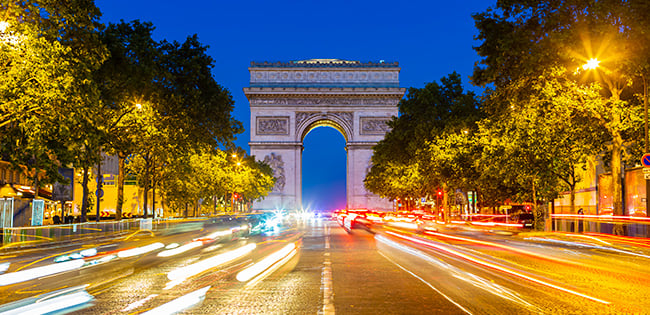 A vibrant nighttime view of the Arc de Triomphe in Paris, France, with bright car light trails creating dynamic streaks on the Champs-Élysées. The monument is illuminated against a deep blue sky, framed by trees and glowing streetlights, creating a lively and iconic scene of the city's elegance and energy.