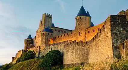 A majestic view of the medieval fortress of Carcassonne in France, bathed in the warm glow of sunset. The castle features towering stone walls, conical blue-topped turrets, and a steep hillside covered in grass and shrubs. The clear evening sky adds to the historical charm and grandeur of this iconic UNESCO World Heritage site.