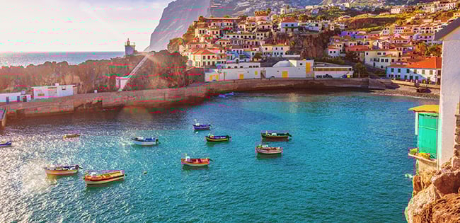 A colorful coastal village built along a hillside overlooks a calm turquoise bay with small boats floating in the water. White buildings with bright red and orange rooftops cluster together near the shoreline under warm evening sunlight.