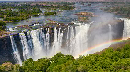 The powerful cascades of Victoria Falls plunge over a cliff into the gorge below, framed by lush greenery and a bright rainbow in the mist.