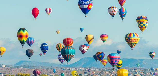 Dozens of colorful hot air balloons float above a sprawling desert cityscape with distant mountains under a clear blue sky.