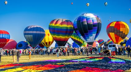 Crowds gather around vibrant hot air balloons in various stages of inflation during a festival, with a bright blue sky overhead.