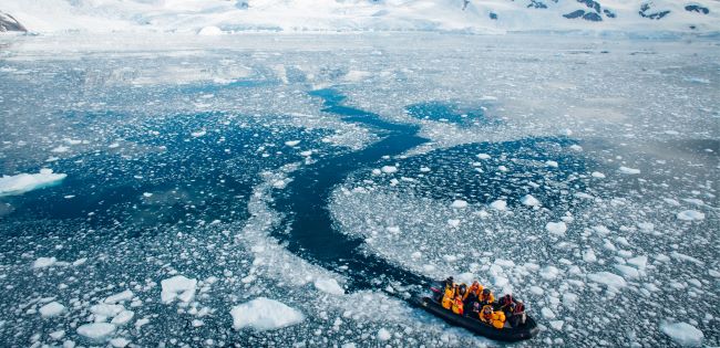A small inflatable boat carrying people in bright orange jackets navigates through icy waters filled with chunks of floating ice, surrounded by a vast, snowy Antarctic landscape.