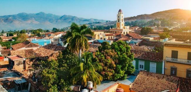 Panoramic view of Trinidad, Cuba, with colonial rooftops, a church tower, lush trees, and distant mountains glowing in the early morning light.