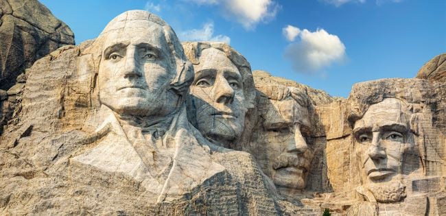 The iconic granite faces of George Washington, Thomas Jefferson, Theodore Roosevelt and Abraham Lincoln carved into Mount Rushmore, set against a blue sky with scattered clouds in the Black Hills of South Dakota.