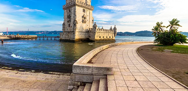 The historic Belém Tower in Lisbon, Portugal, standing at the edge of the Tagus River with a curved stone pathway in the foreground.