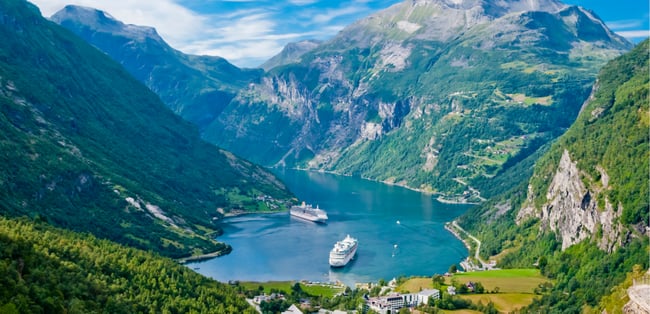 Scenic view of Geirangerfjord in Norway with two cruise ships docked at the fjord, surrounded by steep green mountains and a clear blue sky.