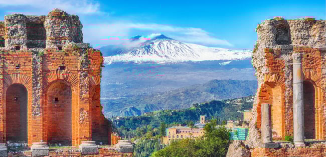 The ruins of the ancient Greek theater in Taormina, Sicily, with its red brick arches and columns framing a spectacular view of Mount Etna in the distance. The snow-capped volcano rises above the lush landscape and pastel-colored buildings below, under a bright blue sky.