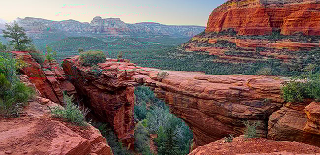 A scenic view of Devil’s Bridge, a natural red rock arch surrounded by Sedona’s iconic rugged cliffs and forested canyon landscape in Arizona.