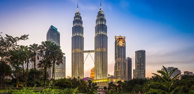 The Petronas Twin Towers in Kuala Lumpur, Malaysia, rise above the skyline at twilight, surrounded by modern high-rises and lush greenery.
