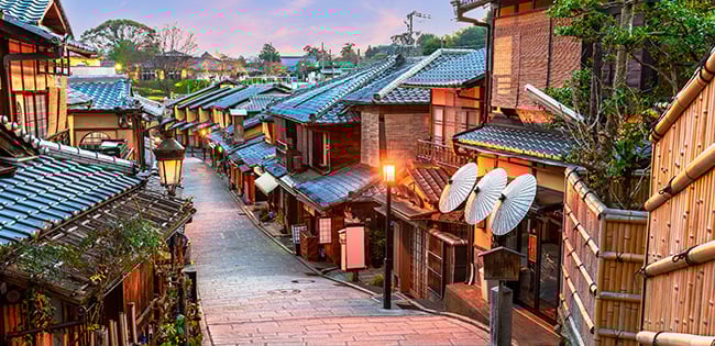 A quiet, lantern-lit street in Kyoto, Japan, lined with traditional wooden buildings and tiled rooftops, just after dusk.