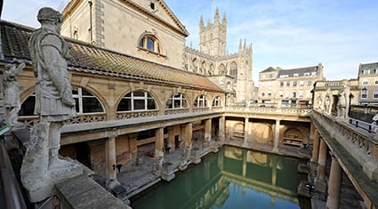 The historic Roman Baths in Bath, England, with arched colonnades, statues, and reflections in the central pool beneath Bath Abbey.