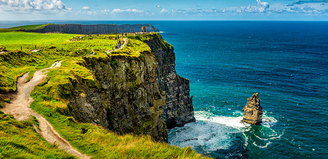 The Cliffs of Moher in Ireland tower above the Atlantic Ocean, with a winding path and grassy fields along the edge of the dramatic cliffs.