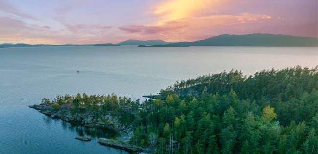 Sunset over the San Juan Islands with a view of forested shoreline, calm waters, and distant mountain silhouettes under a colorful sky.
