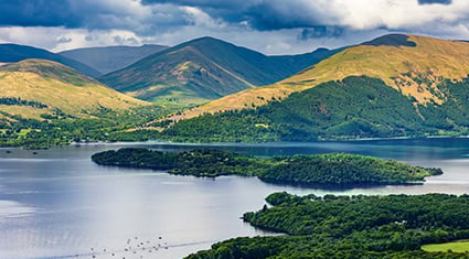 A sweeping view of Loch Lomond in Scotland, with forested islands dotting the water and green hills rolling in the background under a moody sky.