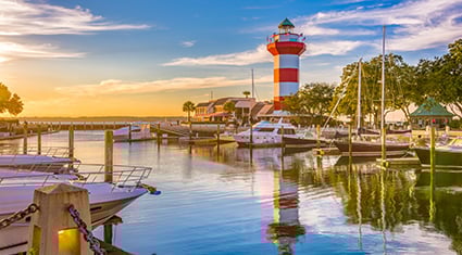 Boats docked at a marina near a red-and-white-striped lighthouse in Hilton Head, South Carolina, at sunset, with calm waters reflecting the sky.