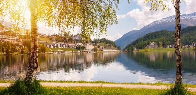 A peaceful lakeside scene in St. Moritz, Switzerland, with the Alps in the background and sunlight filtering through the trees along the shore.