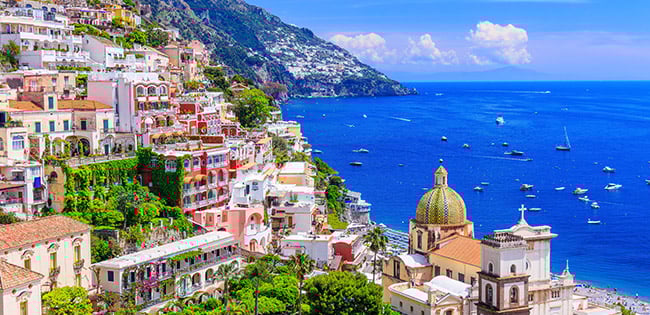 Colorful hillside buildings in Positano, Italy, cascade down toward the deep blue Mediterranean Sea, with boats dotting the water below.