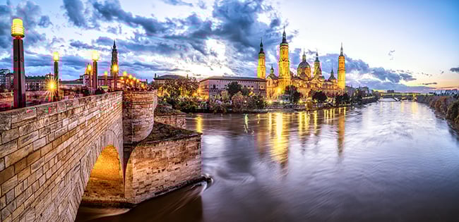 The Basilica of Our Lady of the Pillar in Zaragoza, Spain, lit at twilight and reflecting in the Ebro River, with a stone bridge in the foreground.
