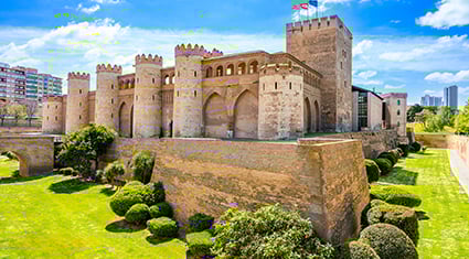 The medieval Aljafería Palace in Zaragoza, Spain, with its distinctive Moorish architecture and manicured green gardens under a bright blue sky.