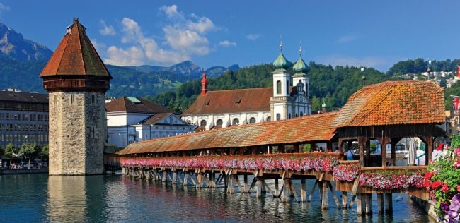 Covered wooden bridge lined with flowers over calm water, with a stone tower and church in the background beneath blue sky and mountains.