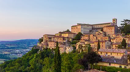 The hilltop town of Trevi, Italy, overlooking the lush Umbrian countryside at sunset, with terracotta buildings nestled among trees.