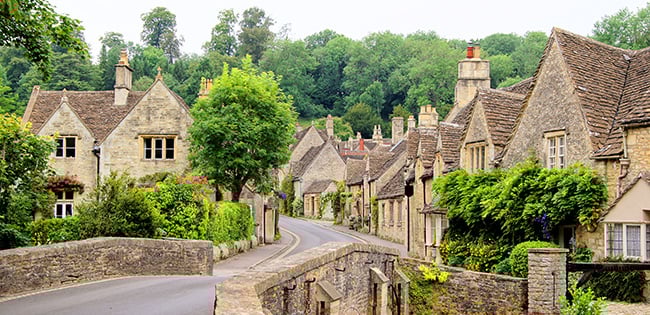 A picturesque street in the Cotswolds, England, lined with charming stone cottages, lush gardens, and a low stone bridge curving through the village.