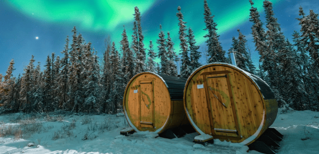 Wooden outdoor saunas at Borealis Basecamp with the aurora borealis stretching across the night sky above a snowy landscape.