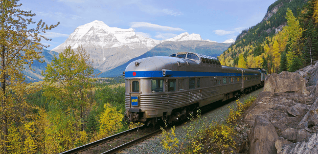 Passenger train traveling along a mountainside track with snow-capped peaks, forested valleys and blue skies in the background.