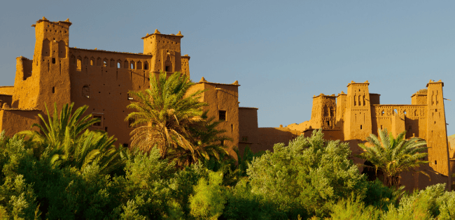 Earthen Moroccan fortress rising above lush palm trees and greenery under a clear blue sky.