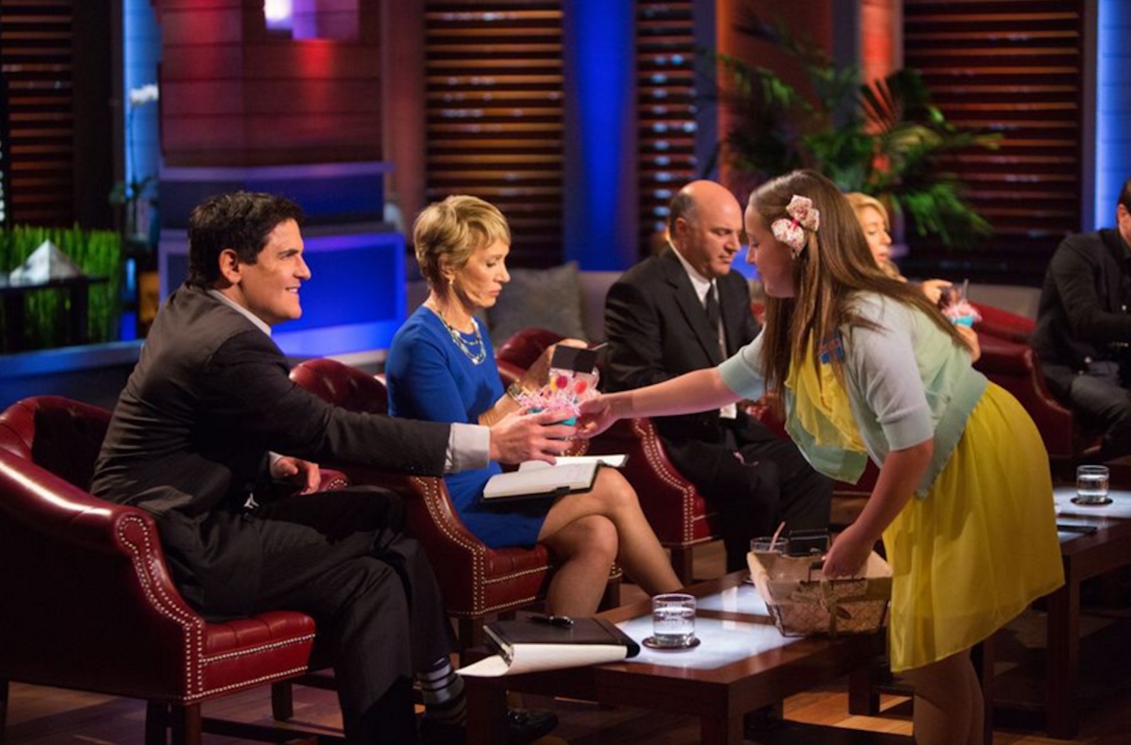 Young founder of a candy company offers samples to Shark Tank investors, leaning forward to hand a product to one of the judges seated in red chairs.