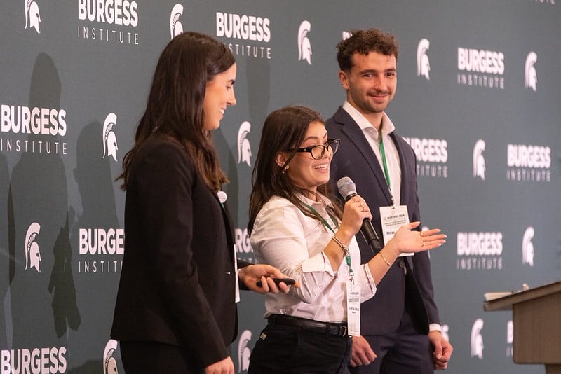 Three students on stage, woman in center holding microphone and speaking.