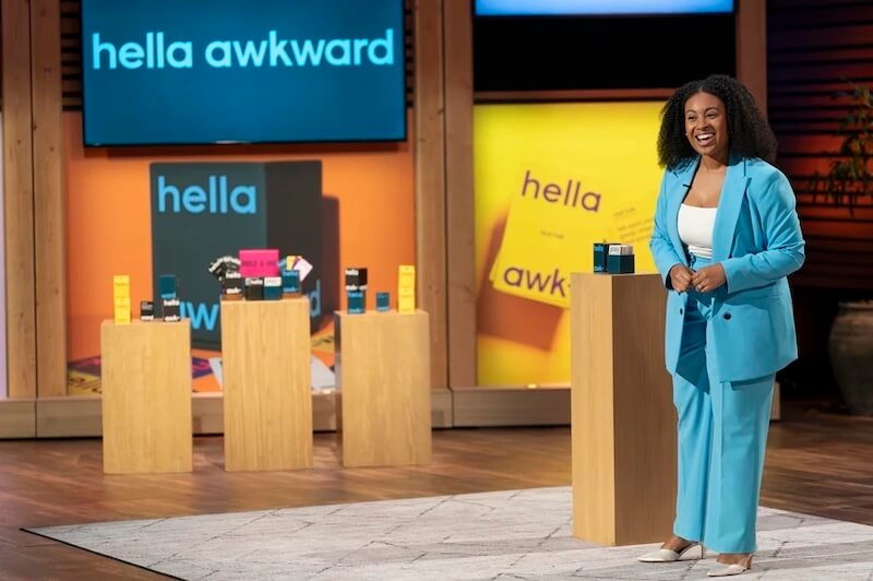Entrepreneur presents Hella Awkward, a social party game, on the Shark Tank stage, standing beside display pedestals with colorful game boxes and branding in the background.