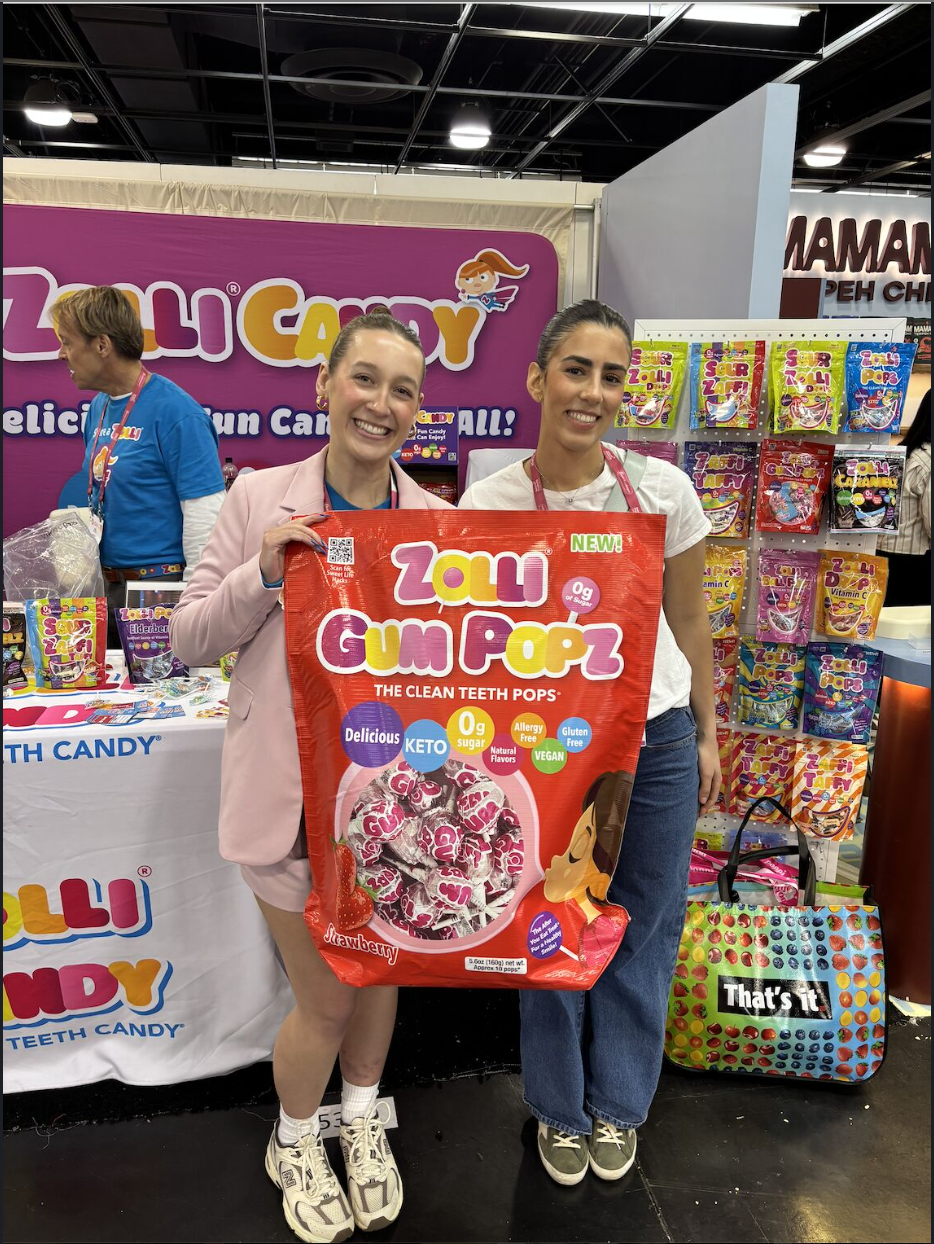 Two women stand at a colorful candy trade show booth, smiling and holding an oversized red bag of “Zolli Gum Popz,” a sugar-free lollipop product. Behind them, a bright pink Zolli Candy display features shelves filled with various colorful candy packages, and a branded table with additional products and promotional materials. One woman wears a light pink blazer and sneakers, while the other wears a white T-shirt and jeans. A man in a blue shirt works at the booth in the background.