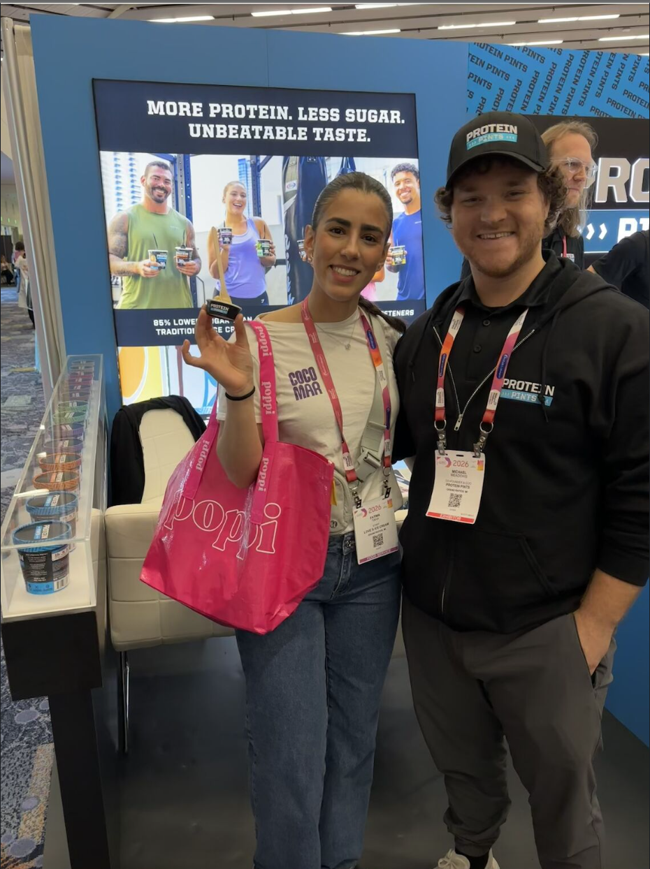 Two people stand smiling at a trade show booth promoting high-protein ice cream. One person holds a small ice cream pint and carries a bright pink Poppi tote bag, while the other wears a black “Protein Pints” hat and hoodie with an event badge. Behind them, a large display screen reads “More protein. Less sugar. Unbeatable taste.” and shows people holding the product, with additional ice cream pints displayed on a shelf at the side of the booth.