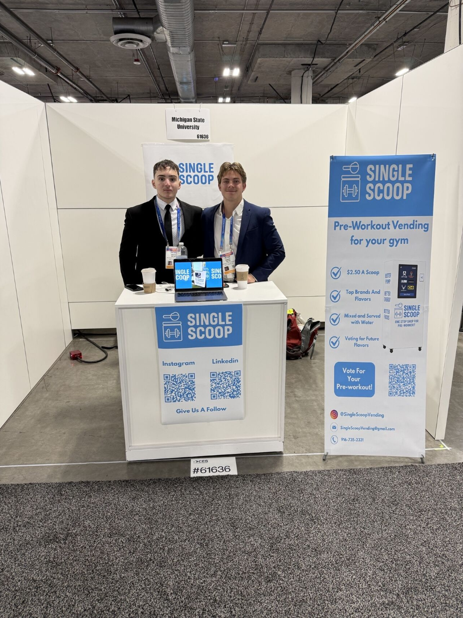 Two young men in suits stand behind a small trade show booth labeled “Michigan State University #61636,” displaying branding for “Single Scoop,” a pre-workout vending concept. The booth features a laptop, coffee cups, QR codes for Instagram and LinkedIn, and a tall banner promoting $2.50 pre-workout scoops, top brands and flavors, and voting for future flavors.