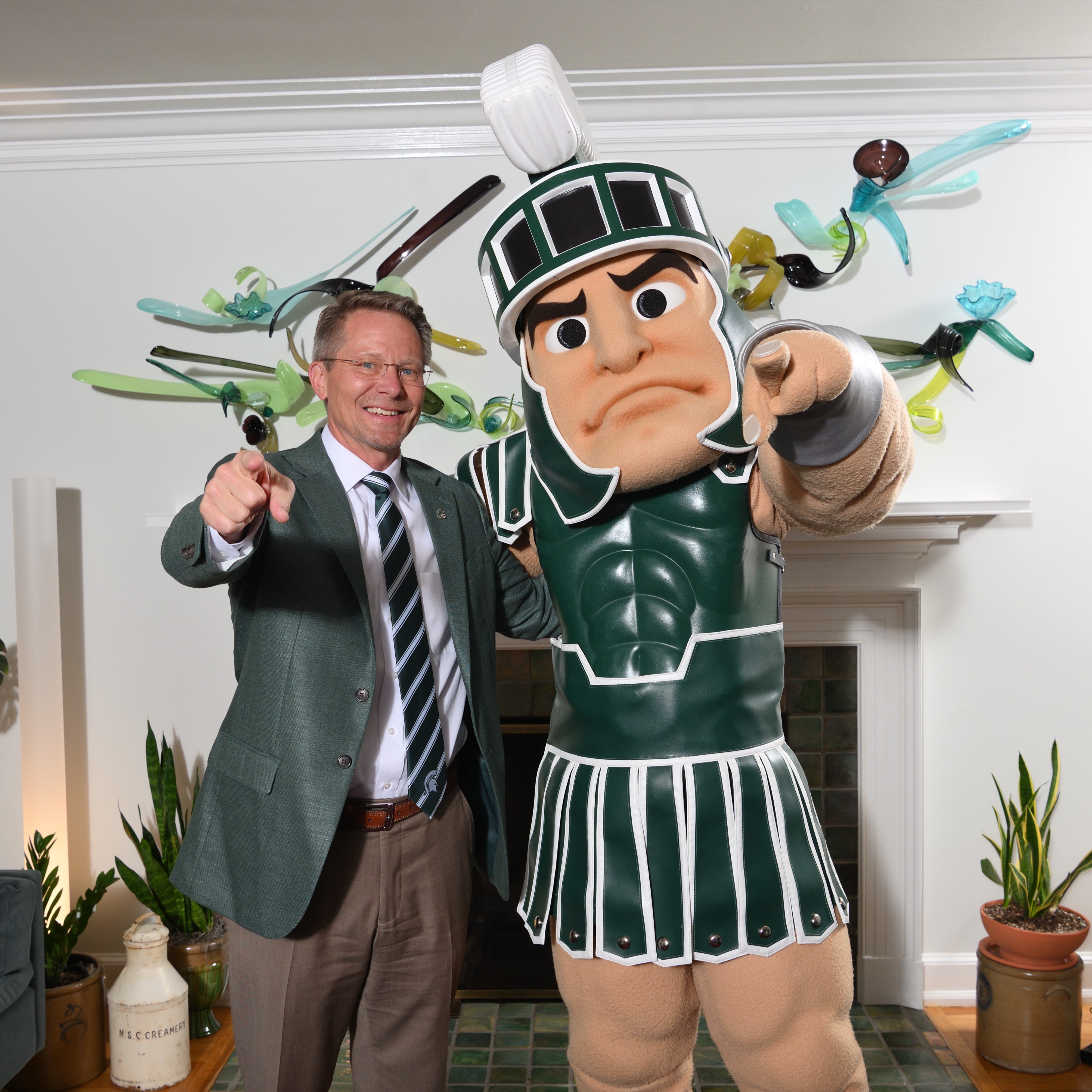 MSU President Kevin Guskiewicz and Sparty standing together at Cowles House while smiling and pointing at the camera