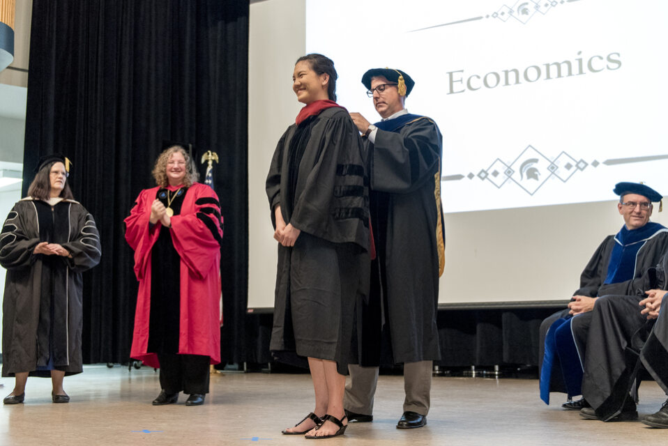 An East Asian female student receives her hood on stage at graduation from the white male department chair while two white female professors applaud. 