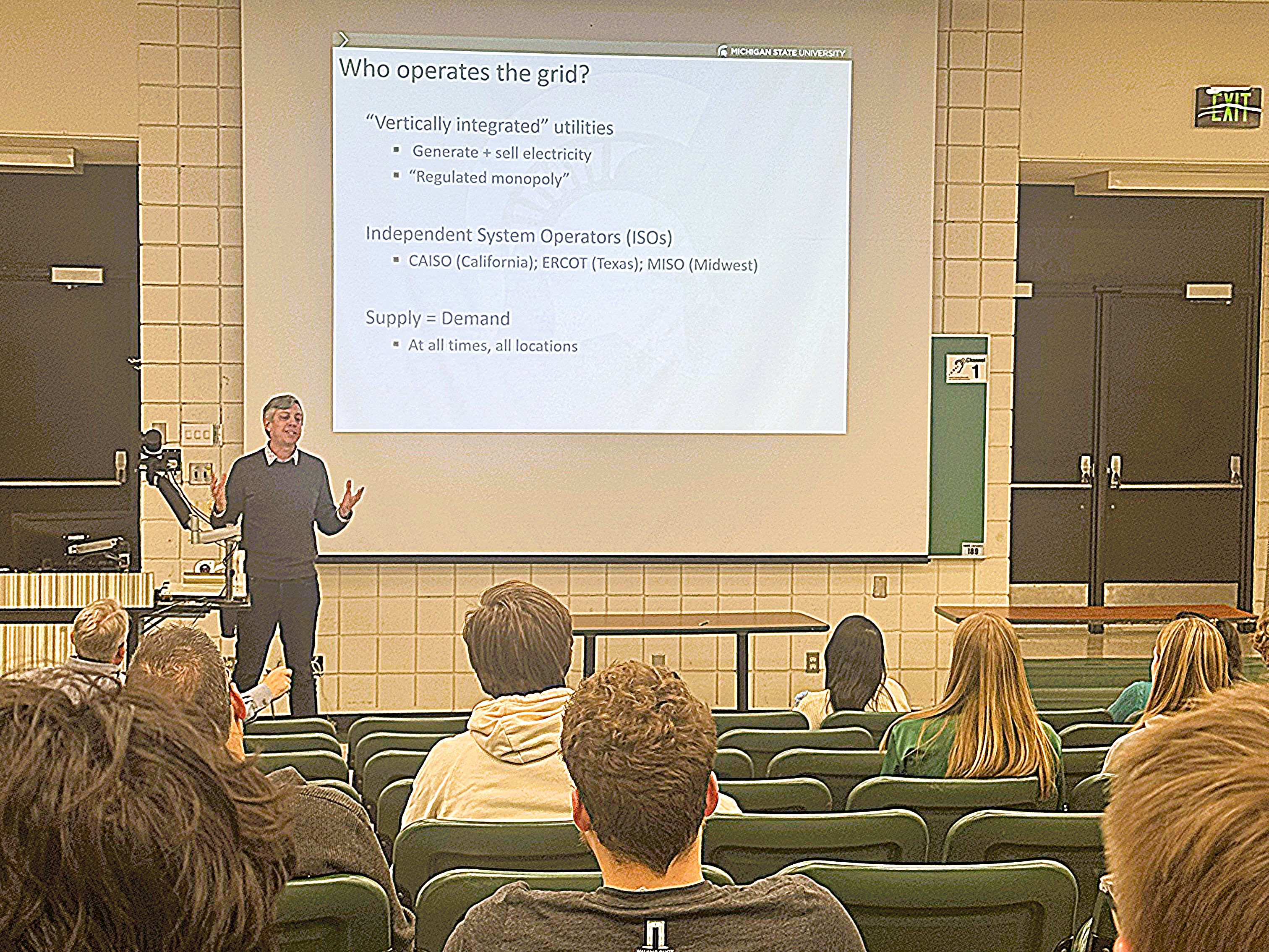 Professor at the front of lecture hall speaking with students.