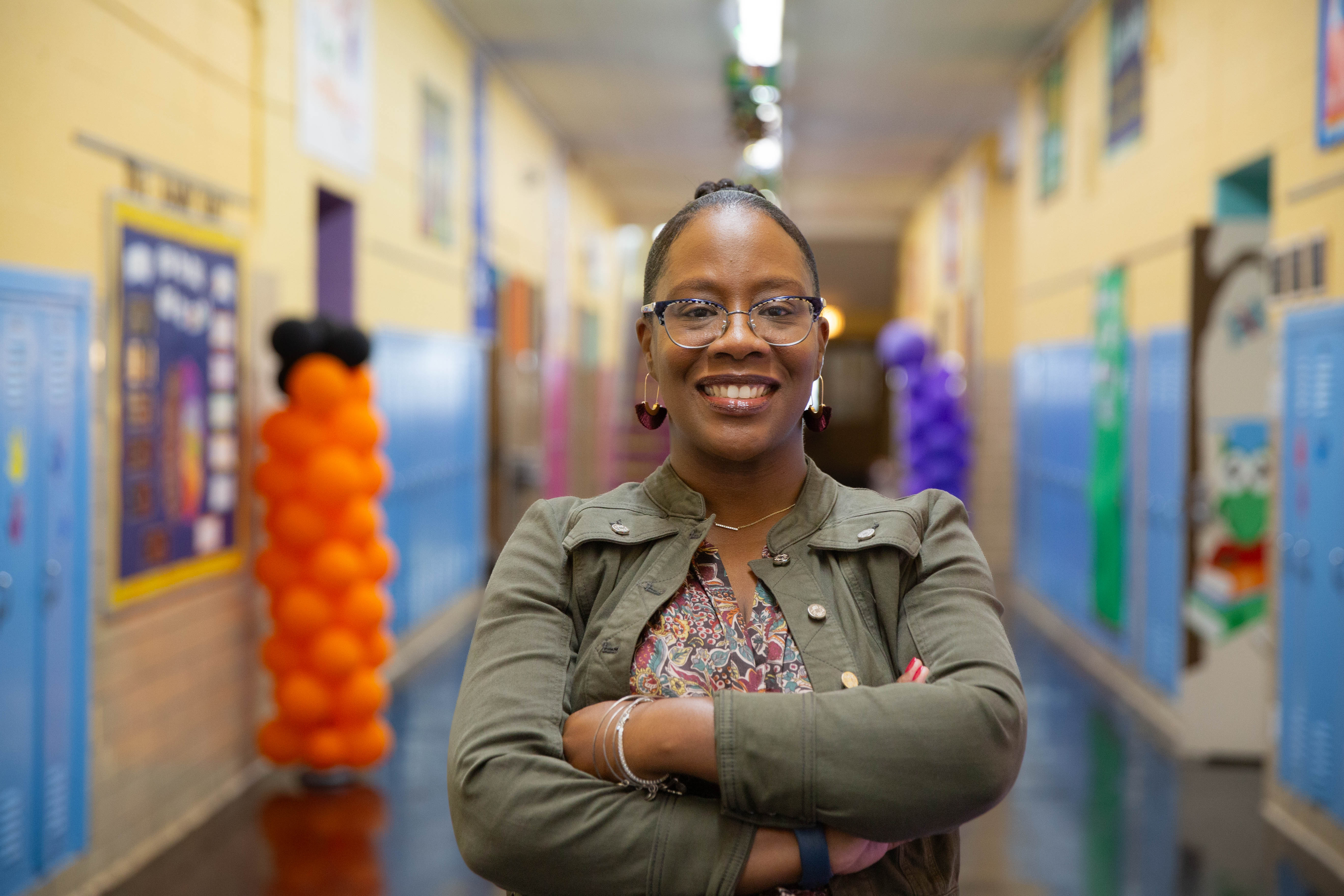Candice Jackson poses for the camera while in a school hallway, smiling and crossing arms across the chest. Candice wears a dark floral dress, a dark green jacket, varying silver accessories and dark-rimmed glasses. Jackson's black hair is pulled back into a bun.