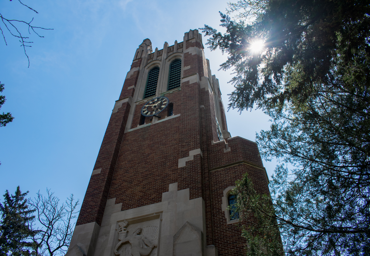 MSU's Beaumont Tower with trees surrounding. The sun is partially hidden behind the trees.