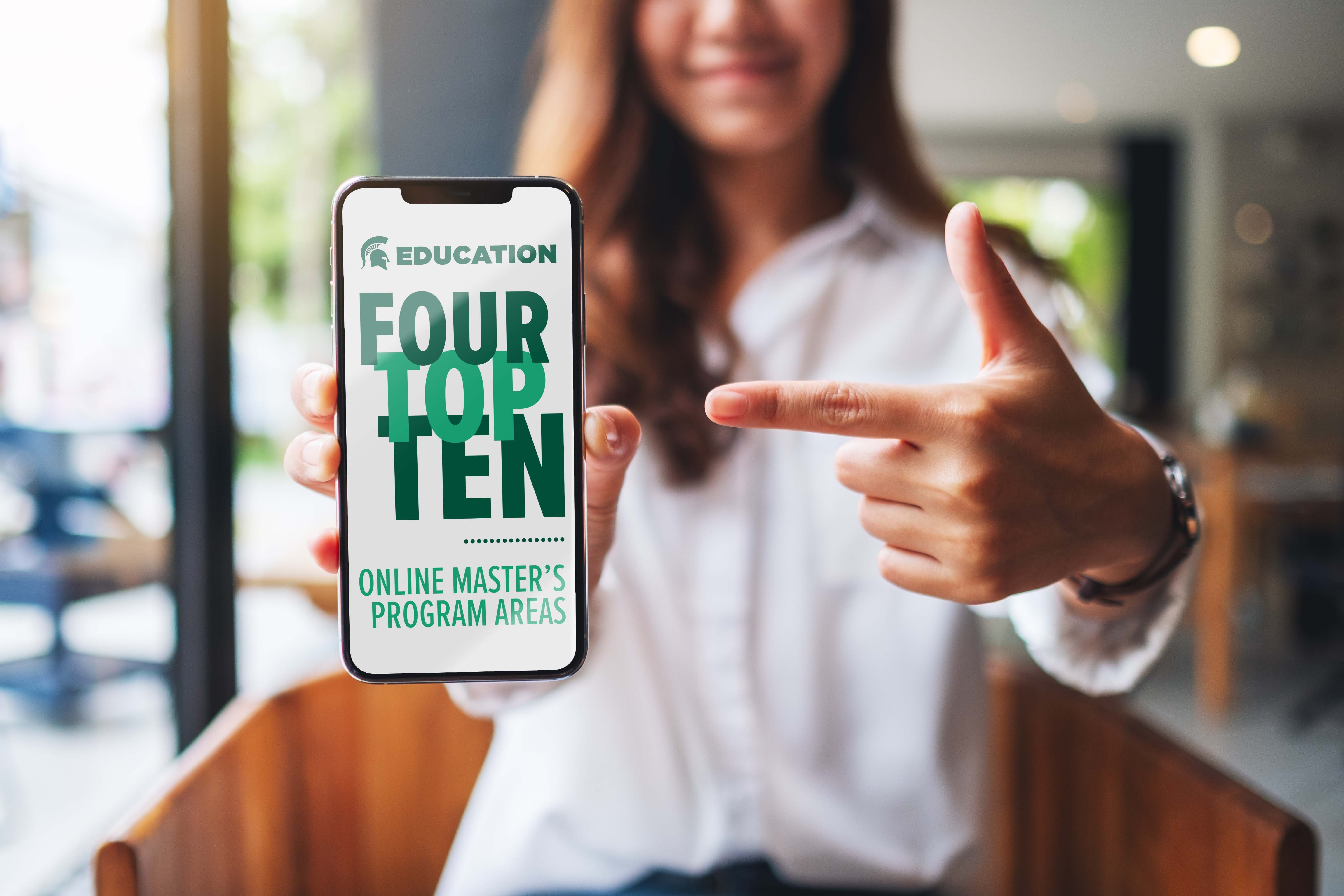 Image of a person holding a phone toward a camera and pointing it to the camera. The phone reads: Four top-10 online master's program areas. 