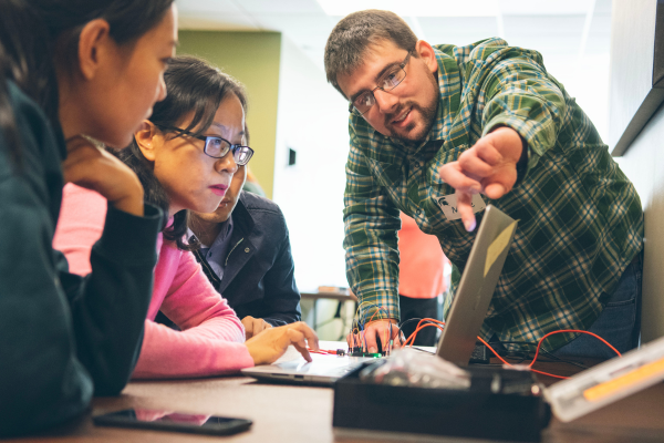 Lachney works with a group of students; Lachney points a finger at a computer while students look on.