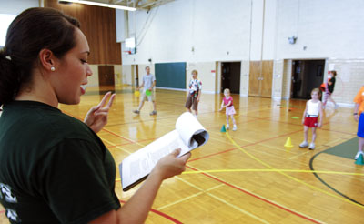 Kinesiology student works with students in a gymnasium. The student holds up a clip board with papers on it and is gesturing to the students, who are standing in rows opposite.