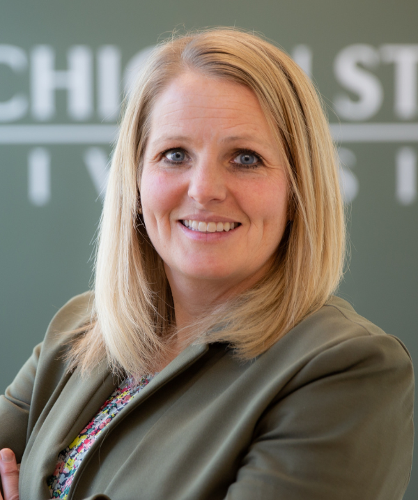 Emily Bouck smiles in front of a sign that reads "Michigan State University." Emily wears a floral shirt under a dark-green blazer. Emily has blonde, shoulder-length hair.