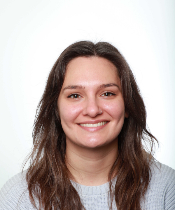 Elizabeth Cloude smiles for the camera against a neutral, white backdrop. Cloude wears a white top. Cloude's brown hair falls past the shoulders.
